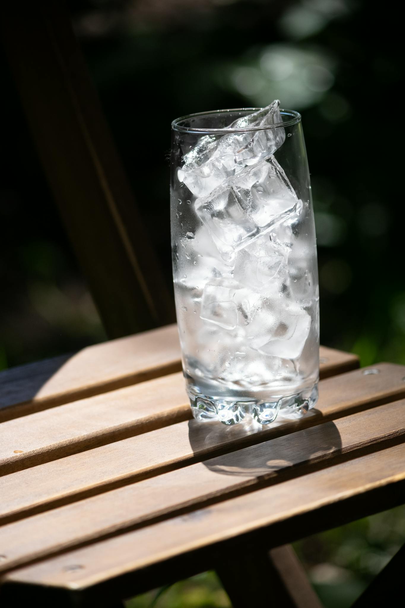 A tall glass filled with ice cubes resting on a wooden table outdoors in sunlight.