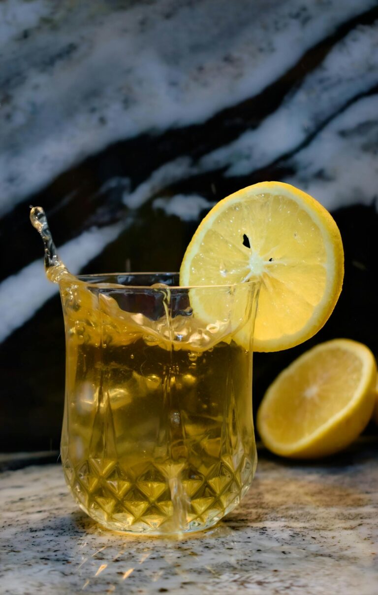 Glass of iced lemon cocktail with a splash, featuring a lemon slice garnish on a marble background.